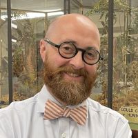 A man with glasses, a beard, and a bowtie stands in front of a glass display case