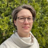 A woman with glasses wearing a cream sweater standing in front of shrubs